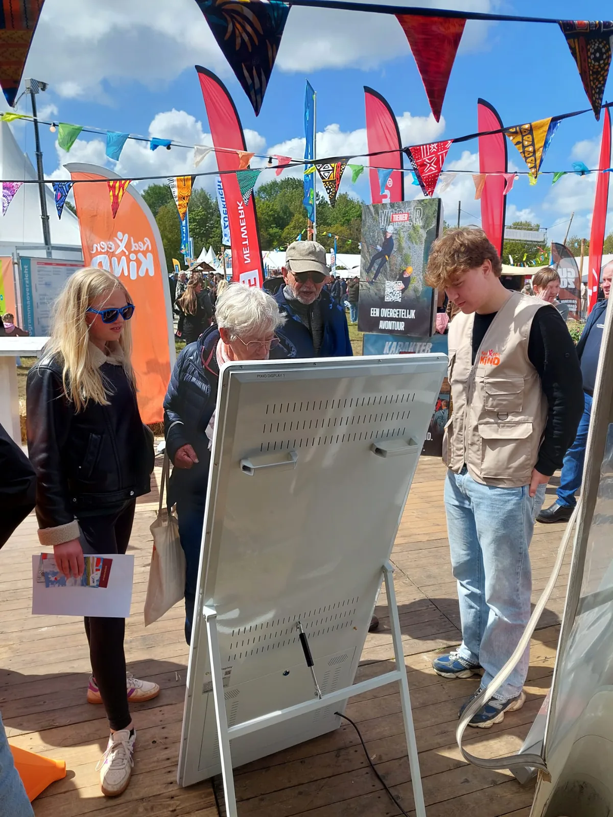 Bezoekers van jong tot oud rond het touchscreen van Red een Kind op een festivalterrein